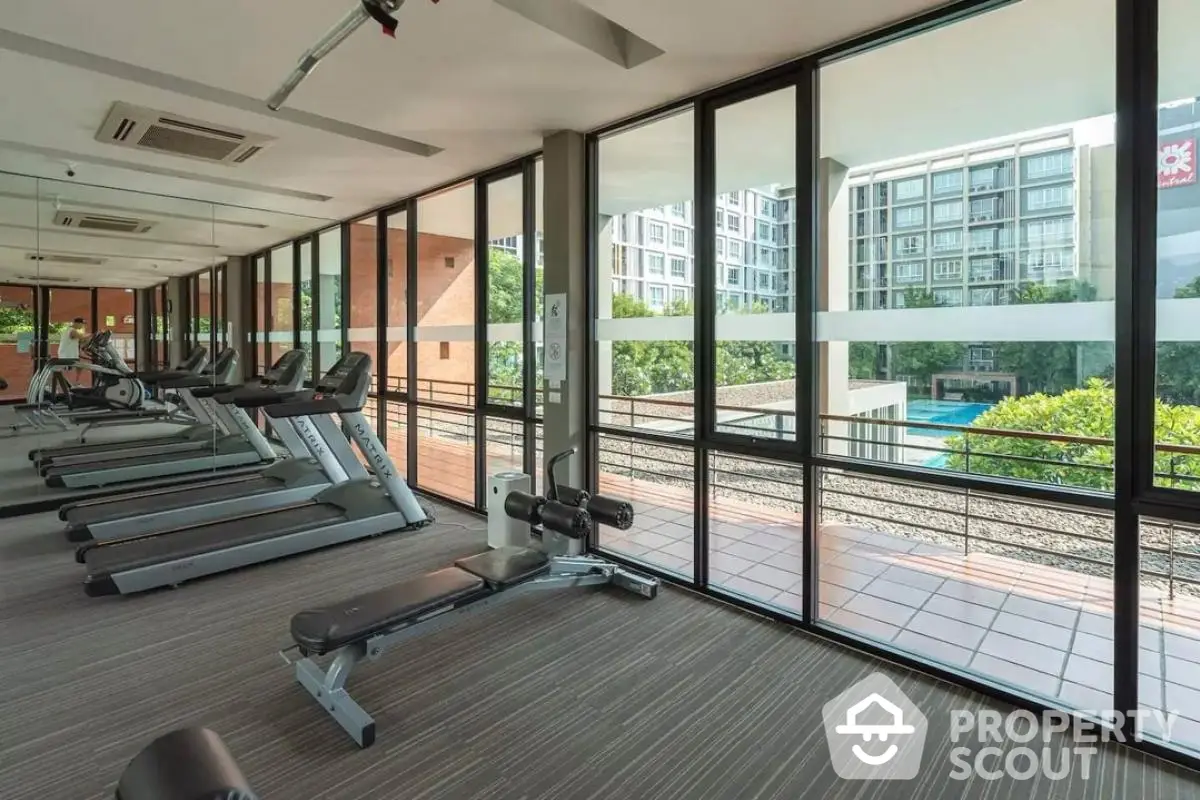 Modern gym with large windows overlooking a pool and greenery in a luxury apartment complex.