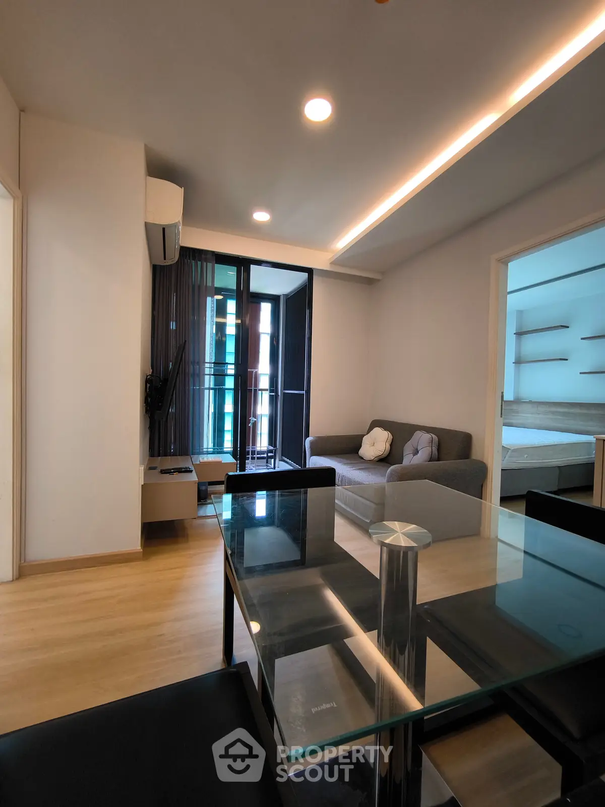 Modern living room with glass dining table and cozy sofa, featuring a view into the bedroom.