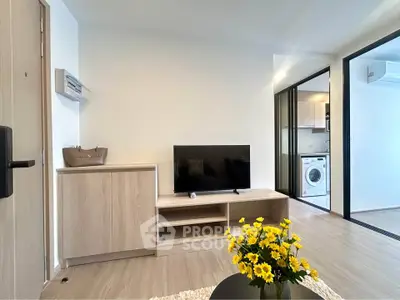 Modern living room with TV, wooden cabinet, and bright flowers, adjacent to a kitchen with washing machine.