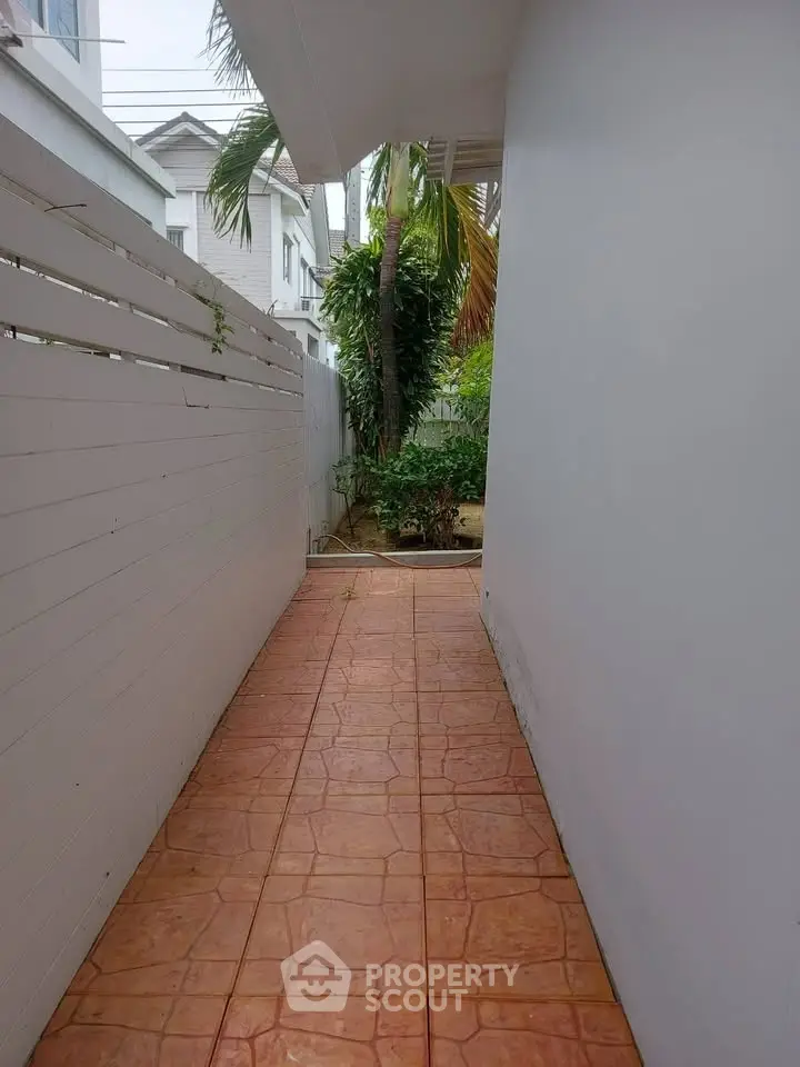 Narrow outdoor walkway with tiled floor and white fence leading to a small garden area.
