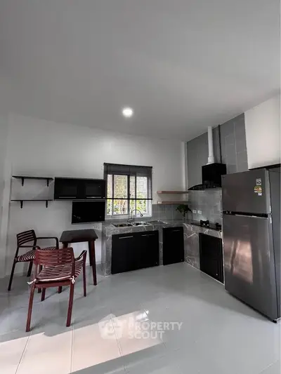 Modern kitchen with sleek black cabinets, stainless steel fridge, and dining area in a bright, open space.