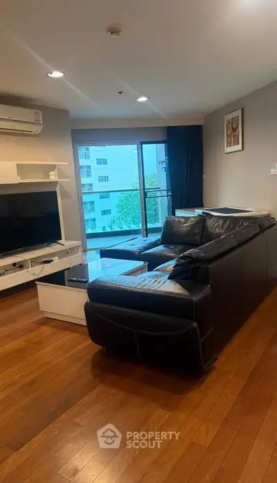 Modern living room with sleek black leather sofa and large TV, featuring wooden flooring and balcony view.