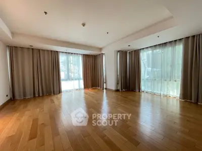 Spacious empty living room with wooden flooring and large windows with curtains.