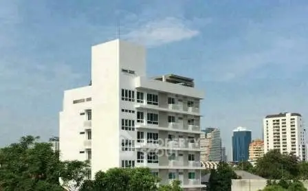 Modern multi-story white building with balconies in urban setting, surrounded by greenery and cityscape views.