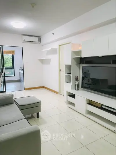 Modern living room with sleek white furniture and large TV, open layout leading to kitchen.