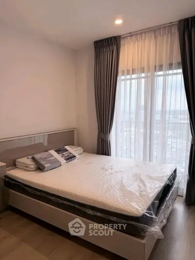 Modern bedroom with large window and stylish curtains, featuring a new mattress and wooden flooring.