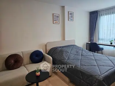 Modern studio apartment with cozy bed, stylish sofa, and natural light from large window.