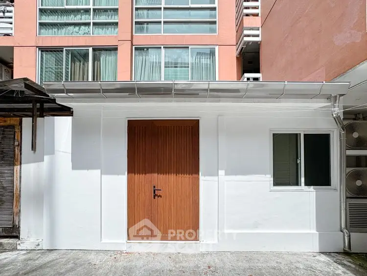 Modern exterior view of a small white building with wooden door and window, adjacent to a larger apartment complex.