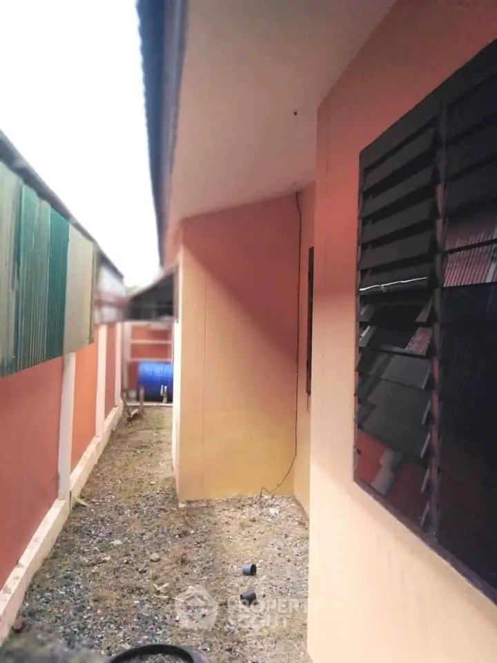Narrow exterior passageway of a residential building with louvered windows and gravel path.