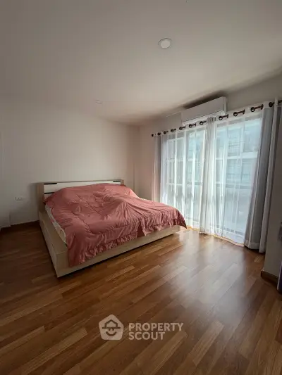 Spacious bedroom with large window and wooden flooring, featuring a cozy bed and ample natural light.