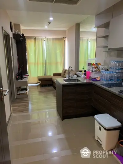 Spacious kitchen with modern cabinetry and glossy tiled flooring opens to a cozy living area, illuminated by natural light from large window with sheer curtains.