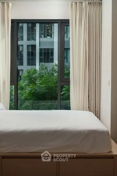 Modern bedroom with large window offering a lush green view, featuring elegant curtains and a cozy bed.