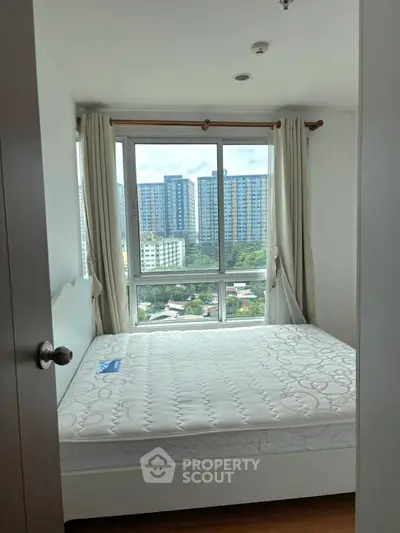 Bright bedroom with large window offering city view, featuring a comfortable bed and elegant curtains.