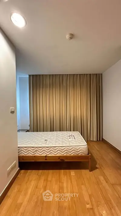 Spacious bedroom with wooden flooring and large window draped with curtains, featuring a minimalist bed frame.
