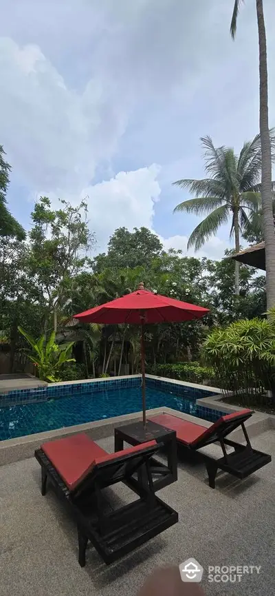 Luxurious outdoor pool area with red sun loungers and umbrella, surrounded by lush greenery and palm trees.