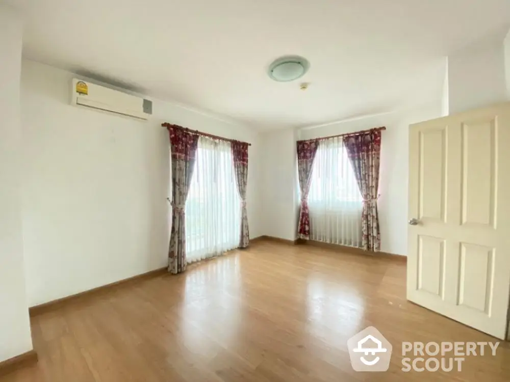 Spacious and well-lit living room with polished wooden flooring, large windows draped with elegant curtains, and a modern air conditioning unit.