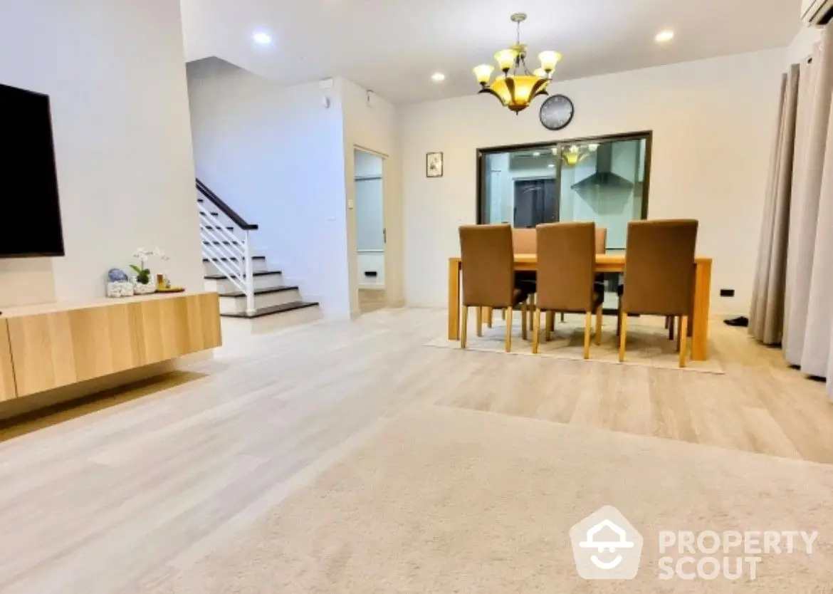 Spacious living room with modern furnishings, elegant dining set, and a sleek staircase leading to the upper level, bathed in natural light from large windows.