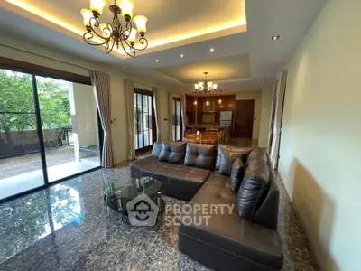 Spacious living room with elegant chandelier and large windows opening to a garden view, featuring a luxurious leather sofa.