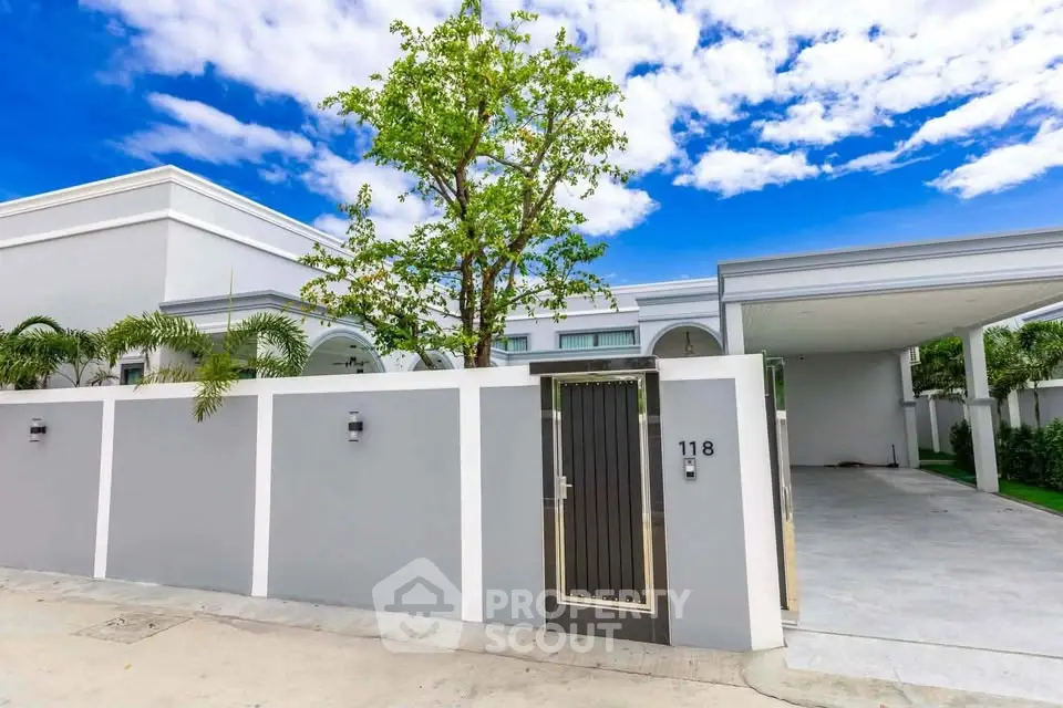 Modern villa exterior with sleek design and spacious driveway, surrounded by lush greenery and clear blue sky.