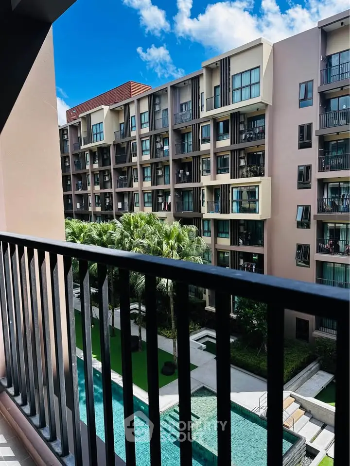 Modern apartment building with pool view from balcony, lush greenery and clear blue sky.
