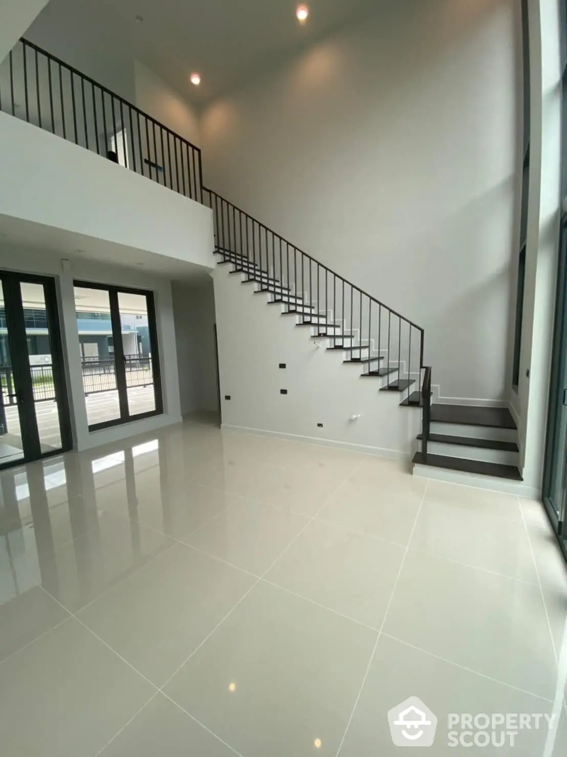 Spacious and modern living area with high ceilings, gleaming white tiles, and an elegant black staircase leading to an upper level, bathed in natural light from large windows.