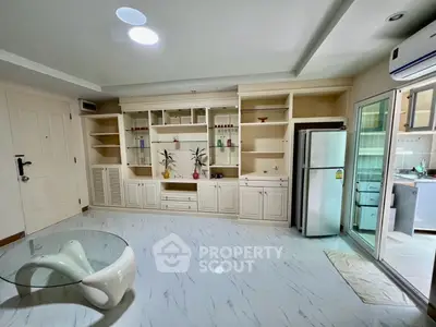 Spacious living room with modern glass table and built-in shelving, adjacent to a sleek kitchen area.
