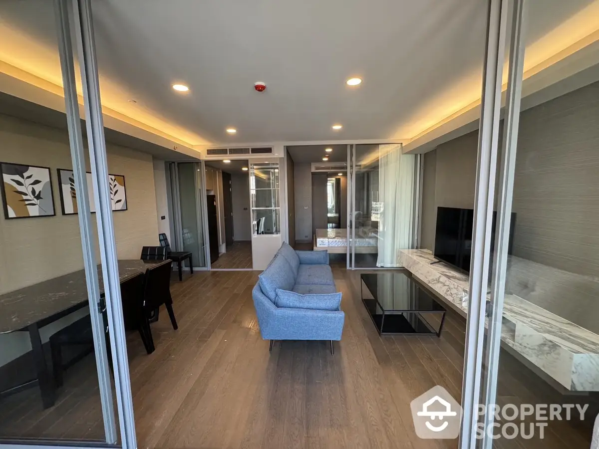 Spacious modern living room with elegant wooden flooring, plush blue sofa, and sleek entertainment unit, leading to a sunlit balcony with a view.