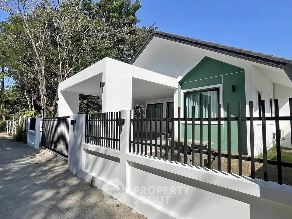 Modern single-story house with sleek design and gated entrance, surrounded by lush greenery.