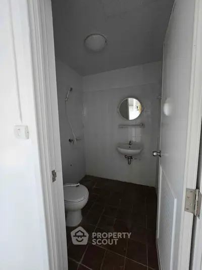 Compact bathroom with white fixtures and tiled floor, featuring a round mirror and shower area.