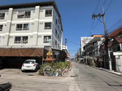 Street view of a residential building with parking and nearby shops under a clear blue sky.