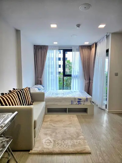 Modern studio apartment with cozy bed and stylish sofa, featuring large windows and elegant curtains.