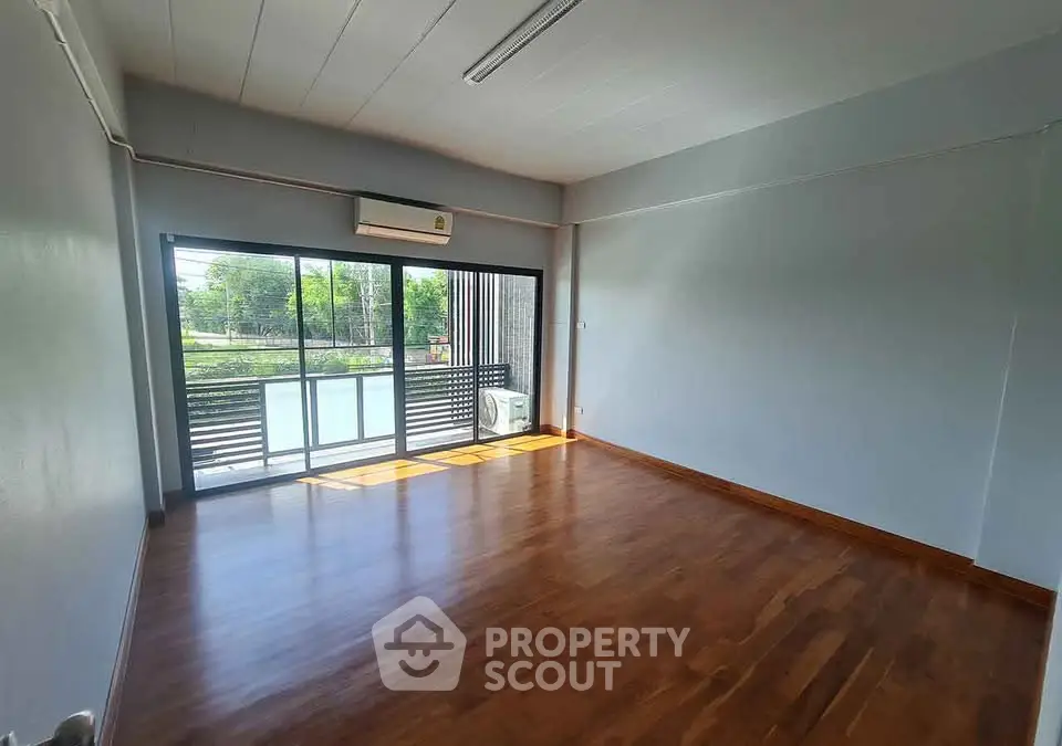 Spacious empty room with wooden flooring and large window overlooking greenery.
