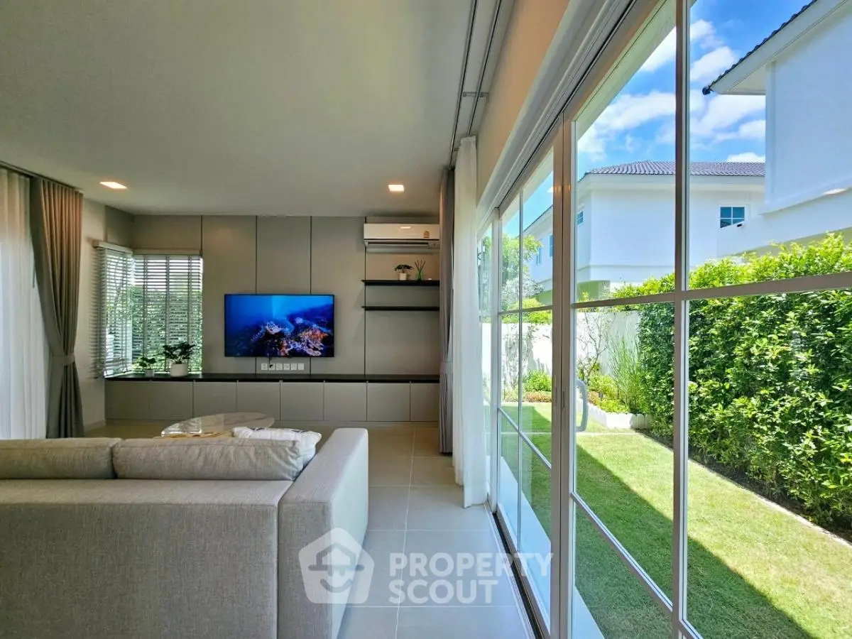 Modern living room with large windows and garden view, featuring sleek furniture and a wall-mounted TV.