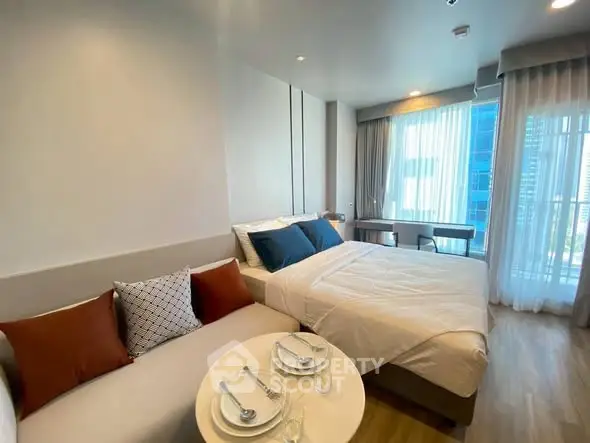 Modern studio apartment with cozy bed and stylish decor, featuring large windows for natural light.
