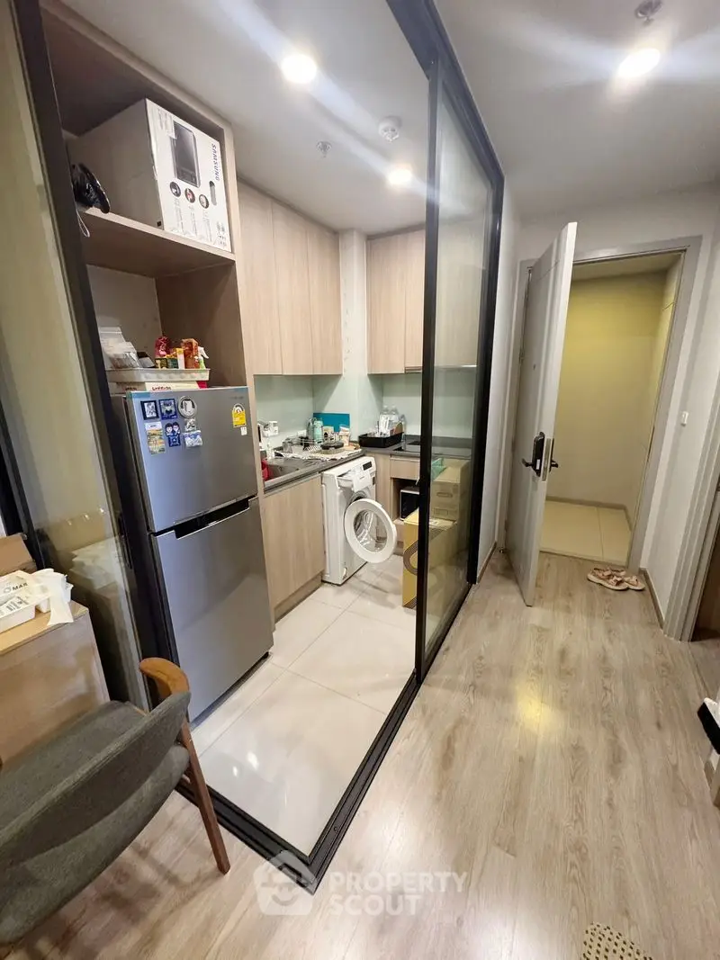 Modern compact kitchen with glass partition, featuring fridge and washing machine.
