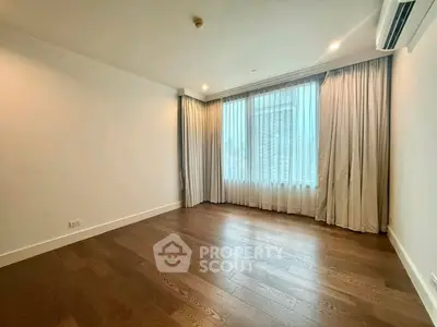 Spacious empty bedroom with wooden flooring and large window with curtains.