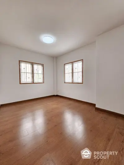 Spacious and well-lit empty room with polished hardwood flooring and dual windows offering ample natural light, ideal for a personalized living space.