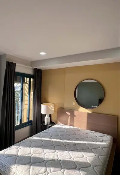 Modern bedroom with natural light, featuring a stylish bed, round mirror, and elegant lamp.