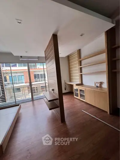 Modern living room with wooden flooring and built-in shelves, featuring large windows for natural light.