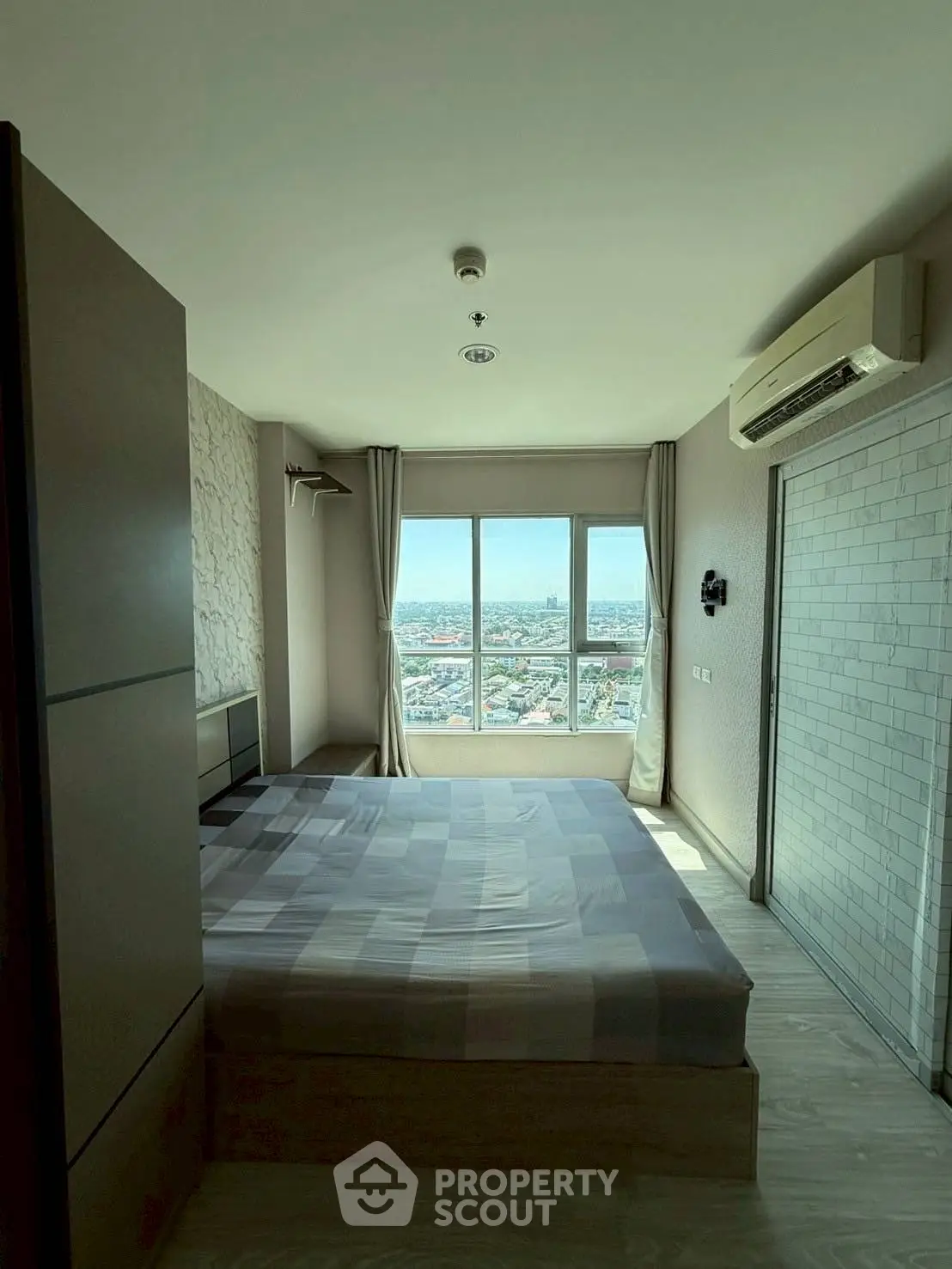 Modern bedroom with city view, featuring large window and air conditioning unit.