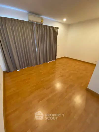 Spacious empty room with wooden flooring and large window with curtains.