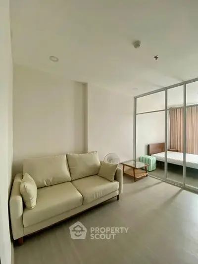 Modern living room with beige sofa and glass partition leading to bedroom in a cozy apartment.