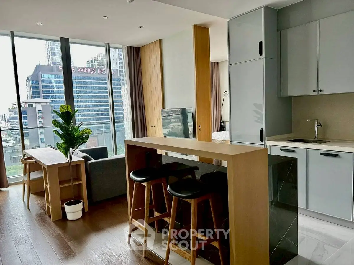 Modern open-plan living room with kitchen and city view, featuring sleek furniture and large windows.