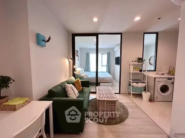 Modern open-plan living room with green sofa and washing machine, leading to a cozy bedroom.
