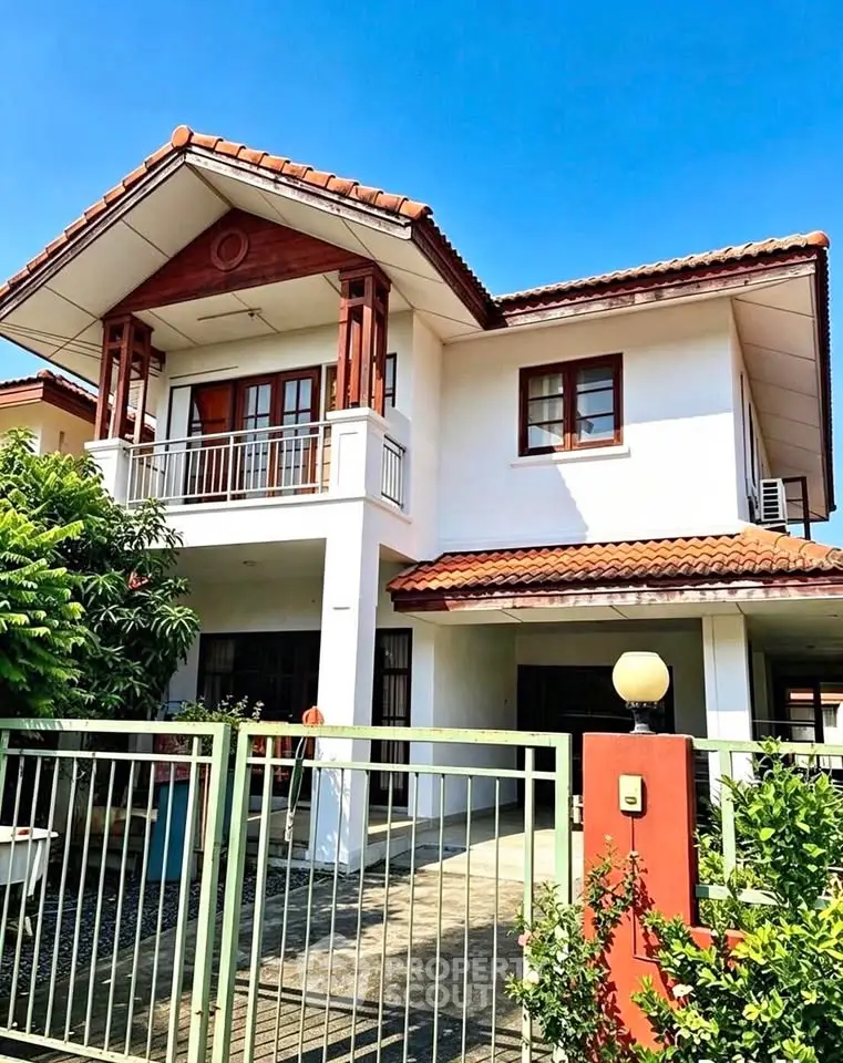 Charming two-story house with a red-tiled roof and spacious balcony, surrounded by lush greenery and a secure gated entrance.