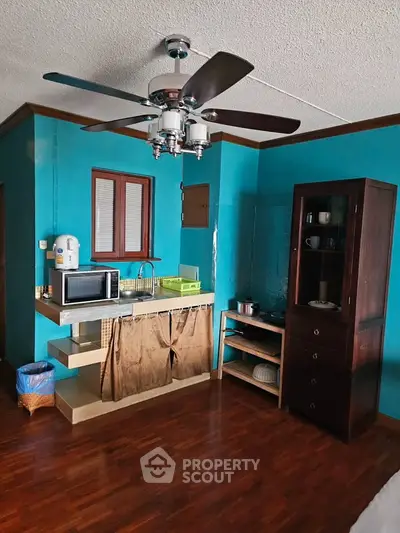 Charming studio with vibrant teal walls, ceiling fan, and compact kitchen area featuring microwave and storage.