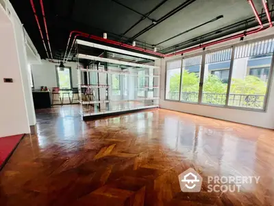 Expansive open-plan living space with polished hardwood floors and abundant natural light, featuring industrial-style red piping and large windows overlooking greenery.