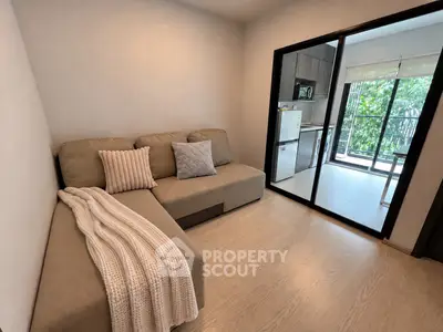 Cozy living room with modern sofa and view into a bright kitchen area with large windows.