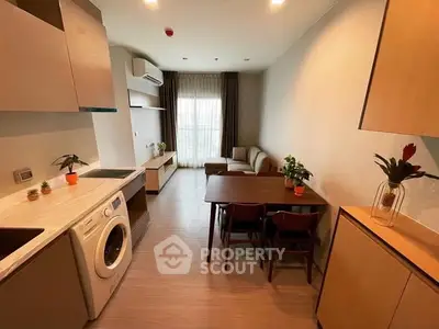 Modern living room with integrated kitchen and washing machine, featuring sleek furniture and natural light.