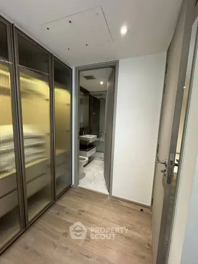 Modern hallway leading to a sleek bathroom with glass shower and wooden flooring.